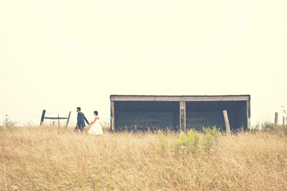 Bride and groom walking through a golden meadow towards a barn